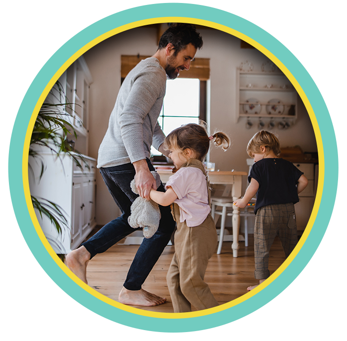Father holding toddler while two children play in a cozy kitchen, framed by a circular border